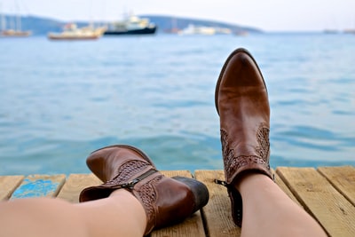 白天，穿着棕色皮革侧拉链靴的人坐在靠近水体的棕色木制码头上